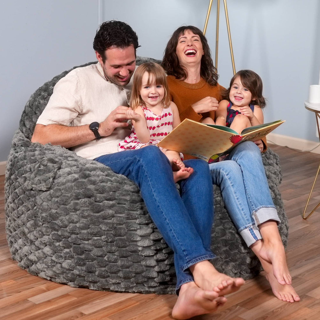 Family of four sitting on a textured gray bean bag chair, reading a book together.