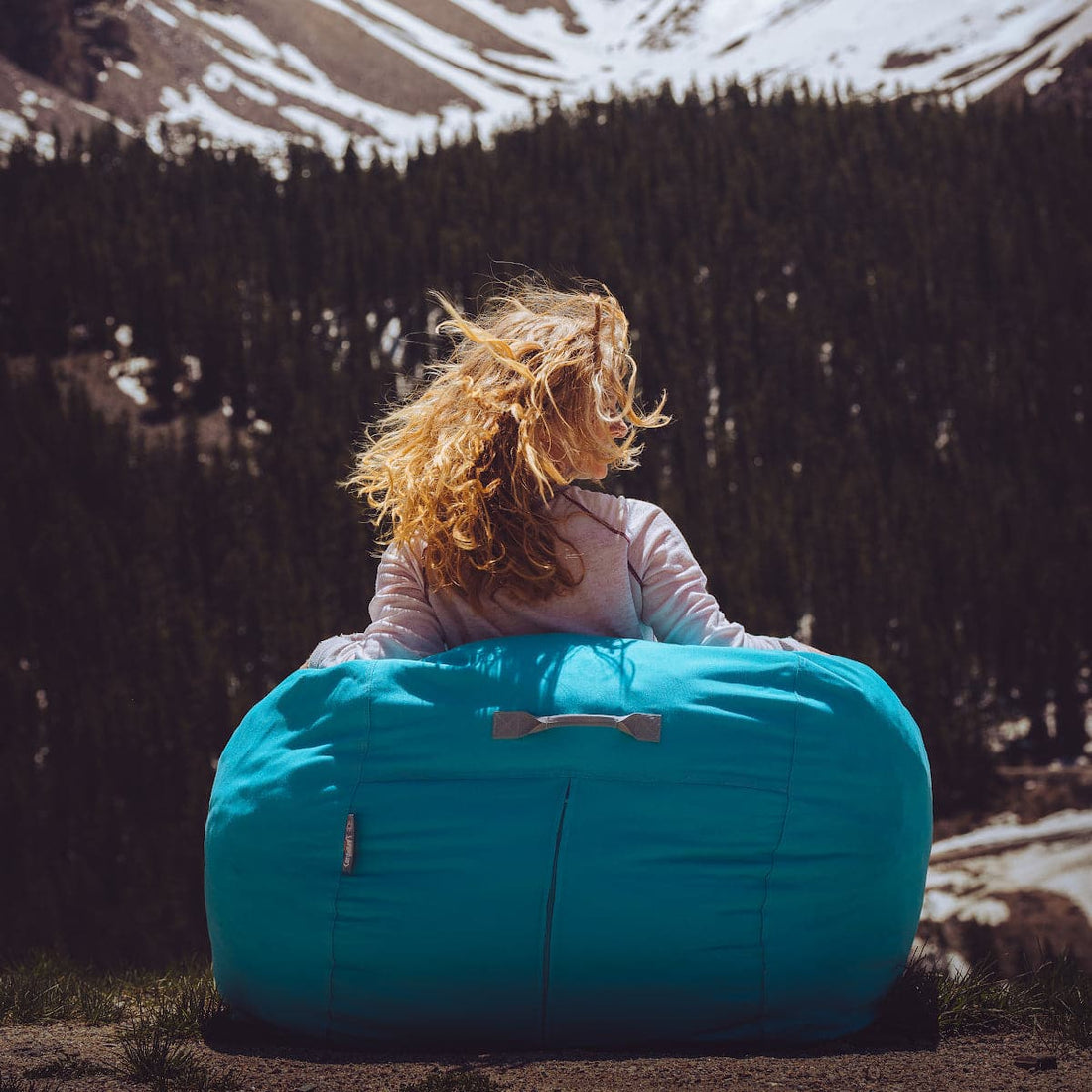 A woman sits in a teal outdoor bean bag in the mountains.