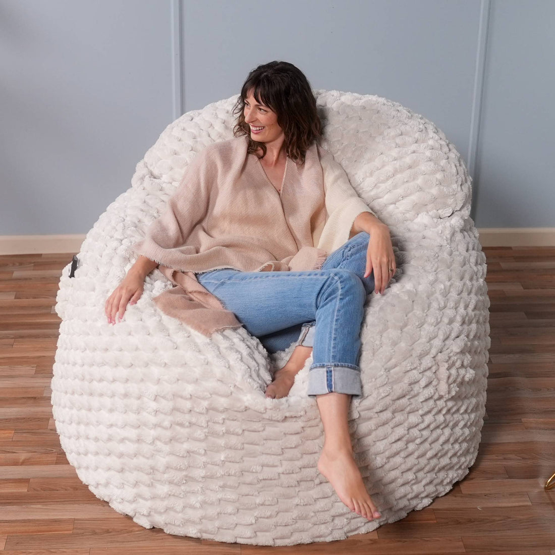 Woman sitting on a textured white bean bag chair in a room with wooden flooring and a gray wall.