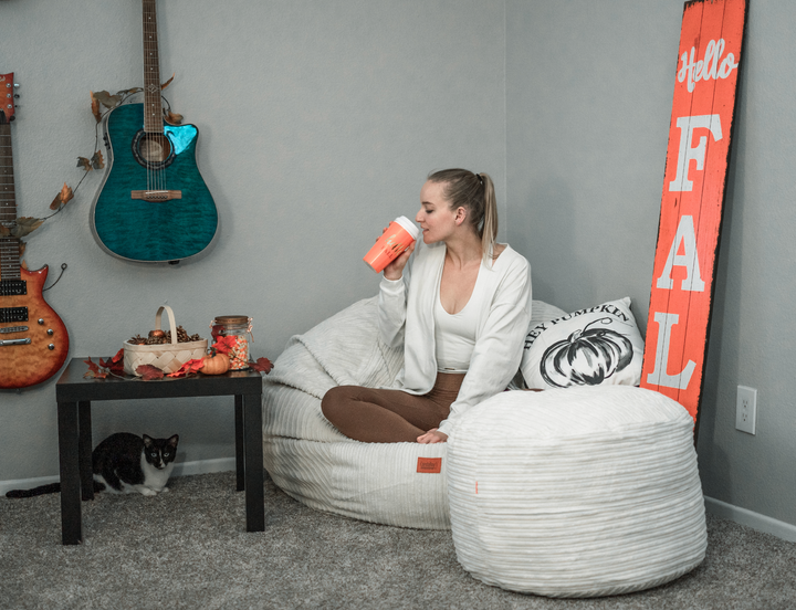 A woman sits on a bean bag chair surrounded by fall decor drinking a fall drink. 