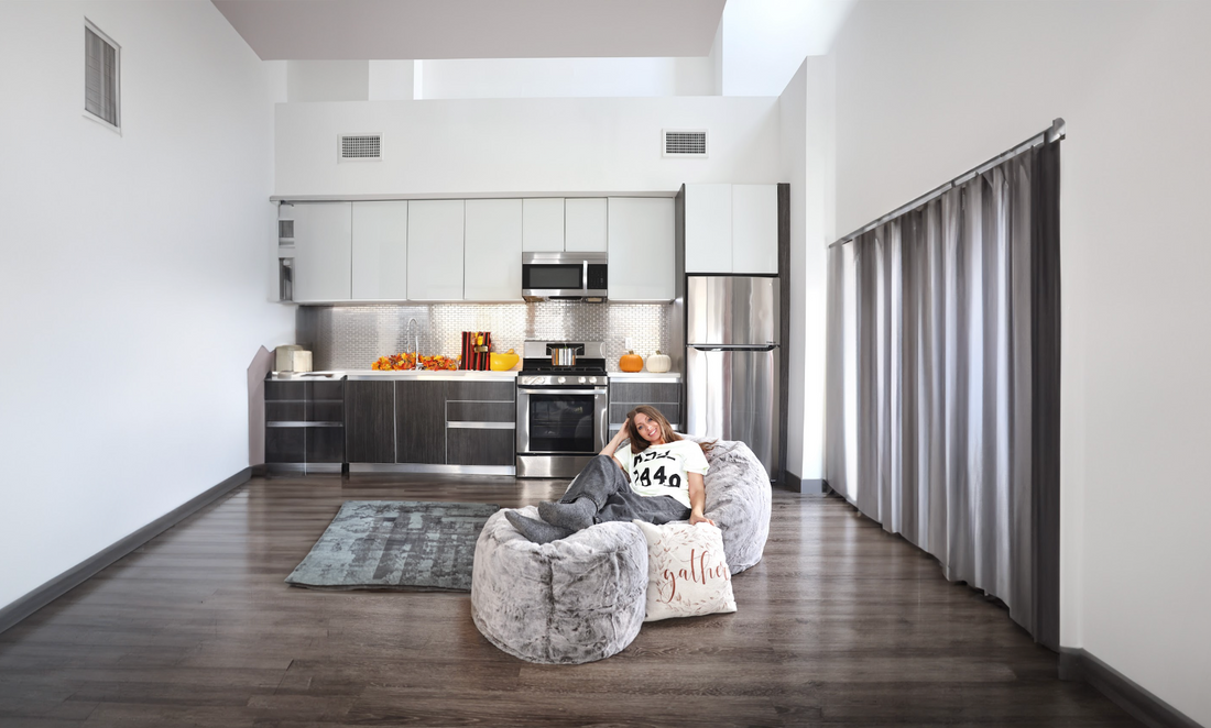 A bean bag chair and pouf footstool are placed in the middle of an open living room with a kitchen in the background. 