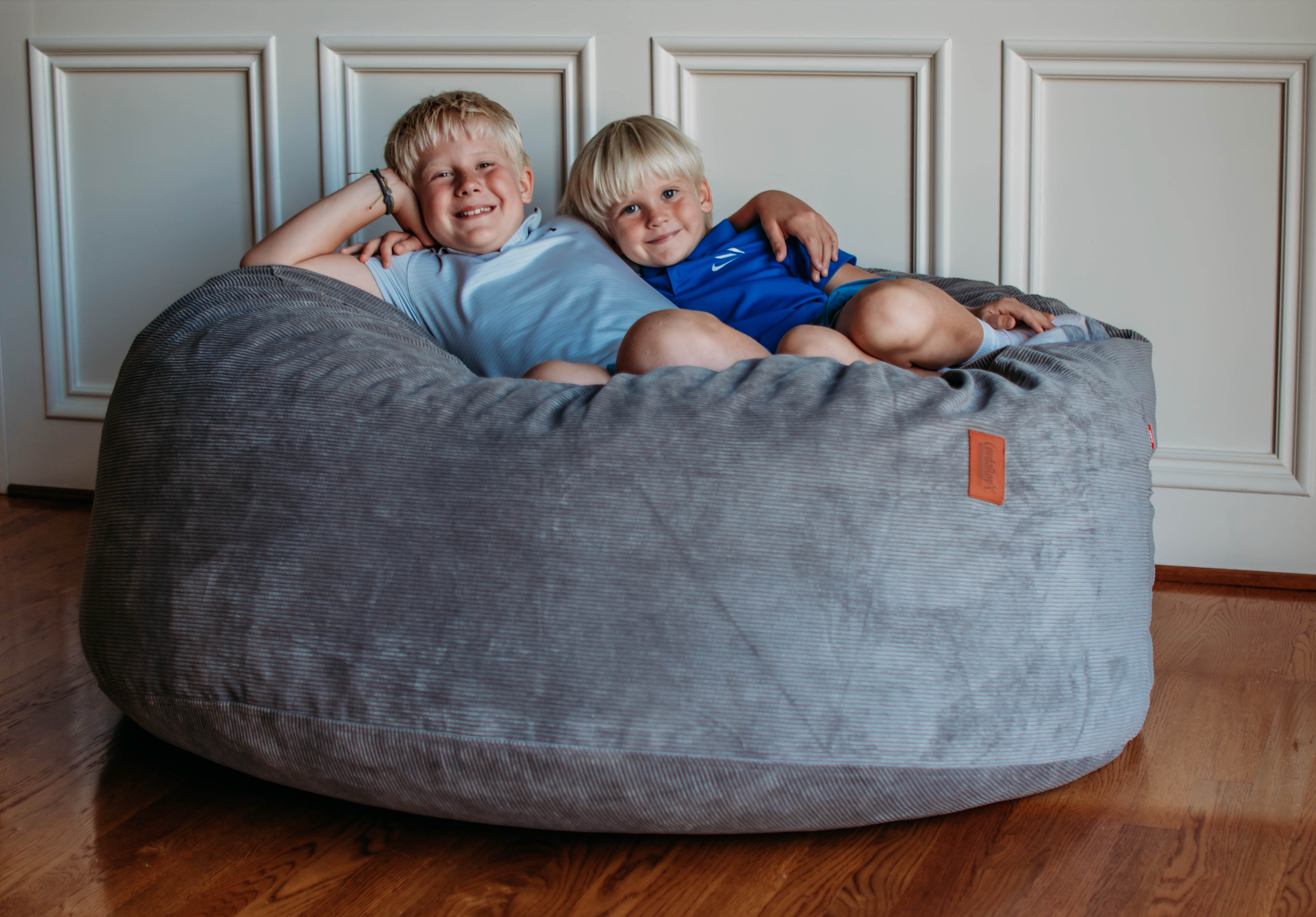 Two kids lay on a cordaroys bean bag chair. 