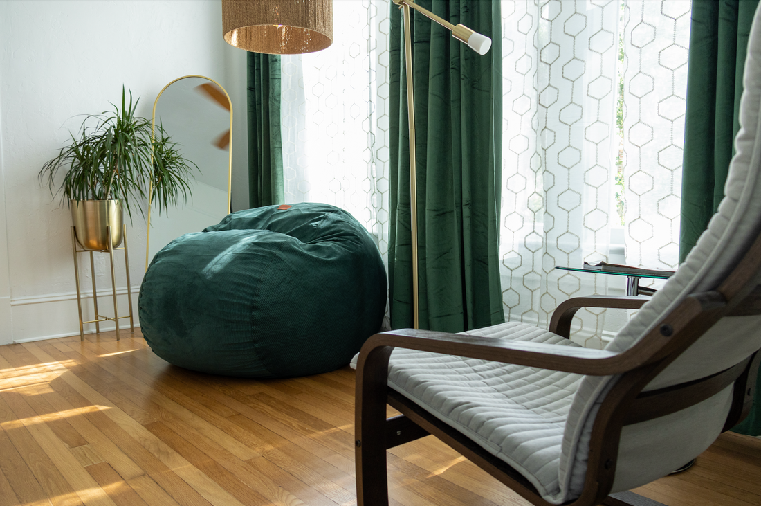 A CordaRoy's Bean Bag sits in the corner of a small room with a chair and lamp. 