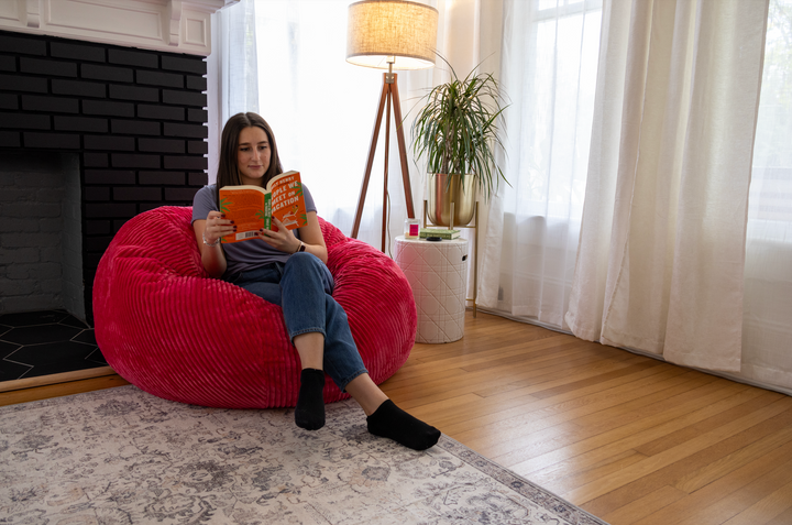 A women reading a book sits on a CordaRoy's round shaped bean bag with a lamp and a plant behind her. 