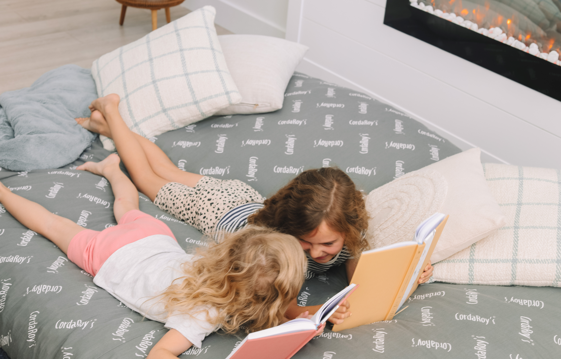 Two girls read books on a cordaroys bean bag bed in front of a fireplace. 