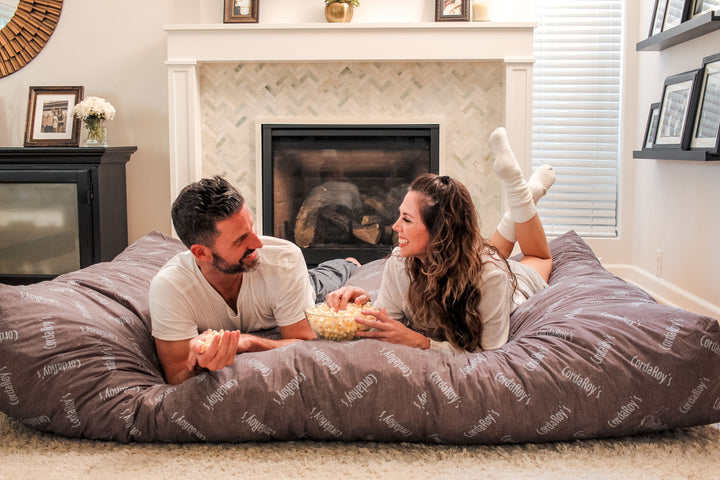 A man and a women lay on a CordaRoy's bean bag eating popcorn. 