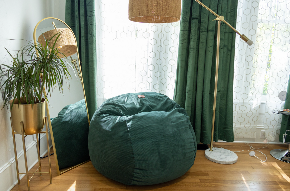 A bean bag chair is in an apartment surrounded by a light, mirror, plants, and curtains covering a window.