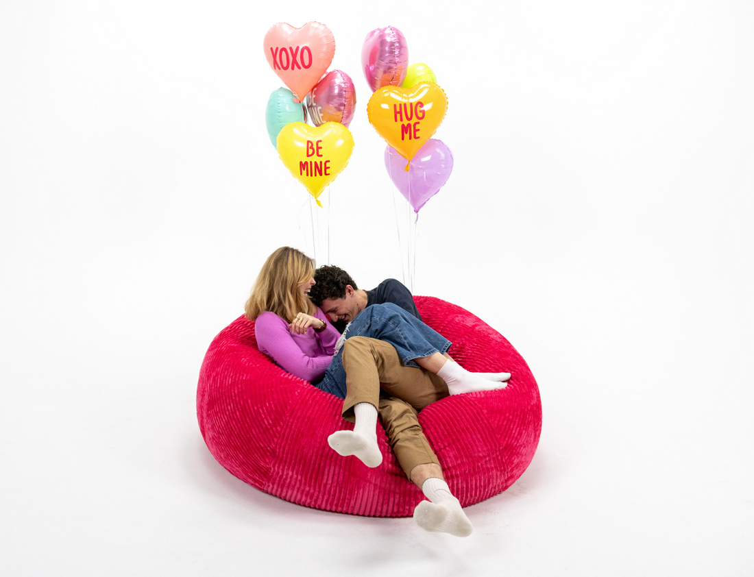 A couple sitting on a red CordaRoy's bean bag chair with heart shaped balloons in the background