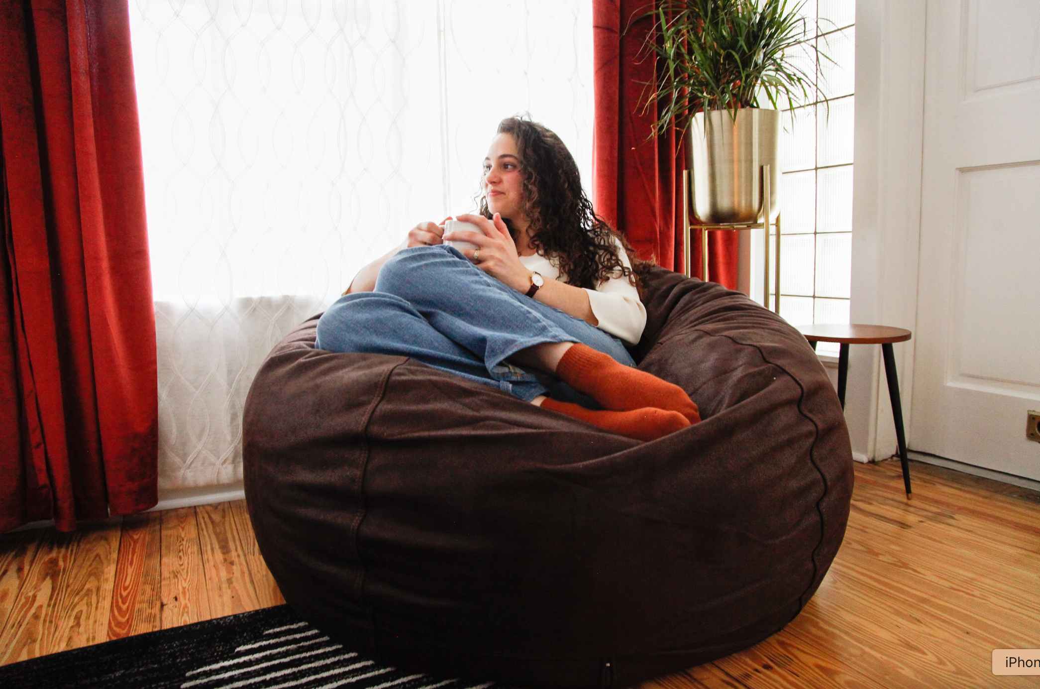 A woman sits on a cordaroys bean bag chair in a room lit by sun.