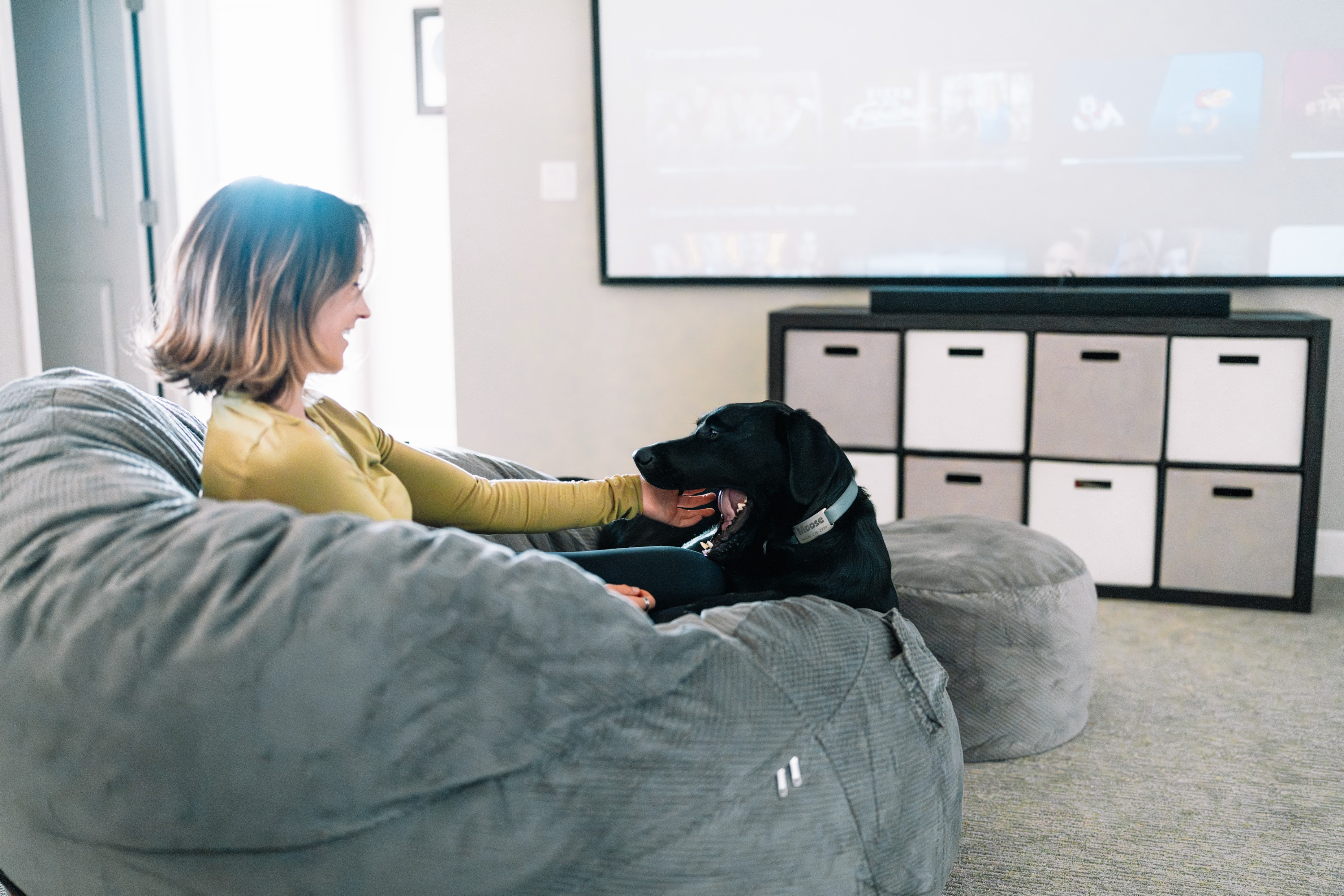A women sits on a cordaroys with her dog watching a large TV.