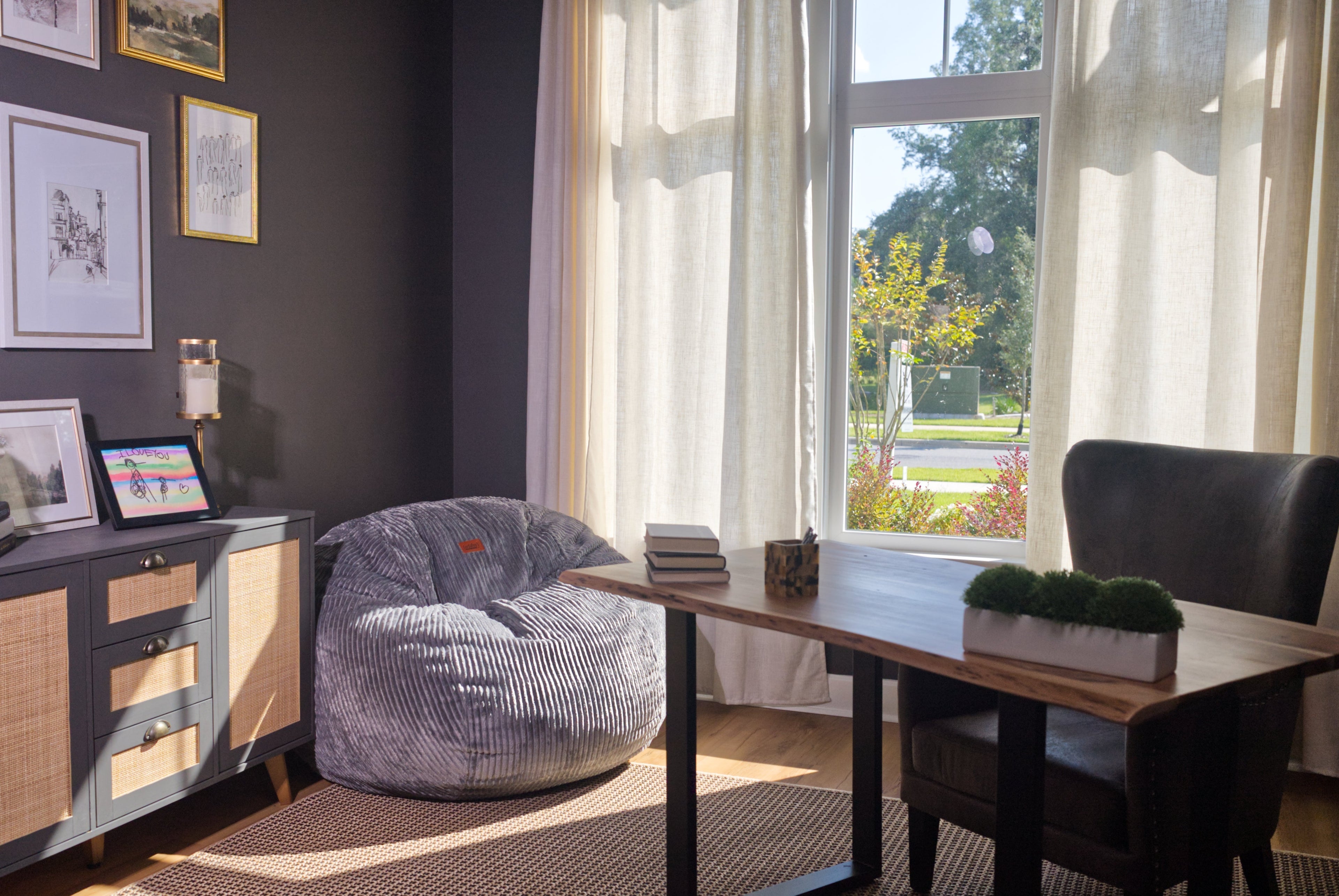 A cordaroys grey bean bag chair is placed in the corner of an office by the window. 