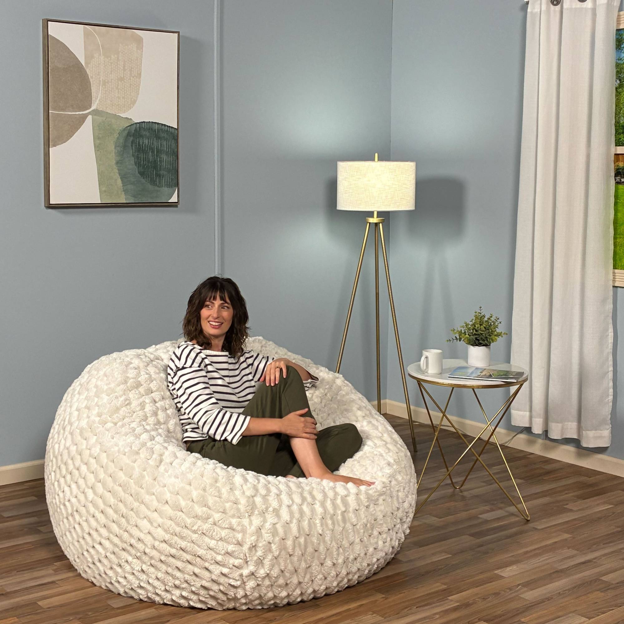 Woman sitting on a textured white bean bag chair in a room with a lamp, small table, and decorative elements.