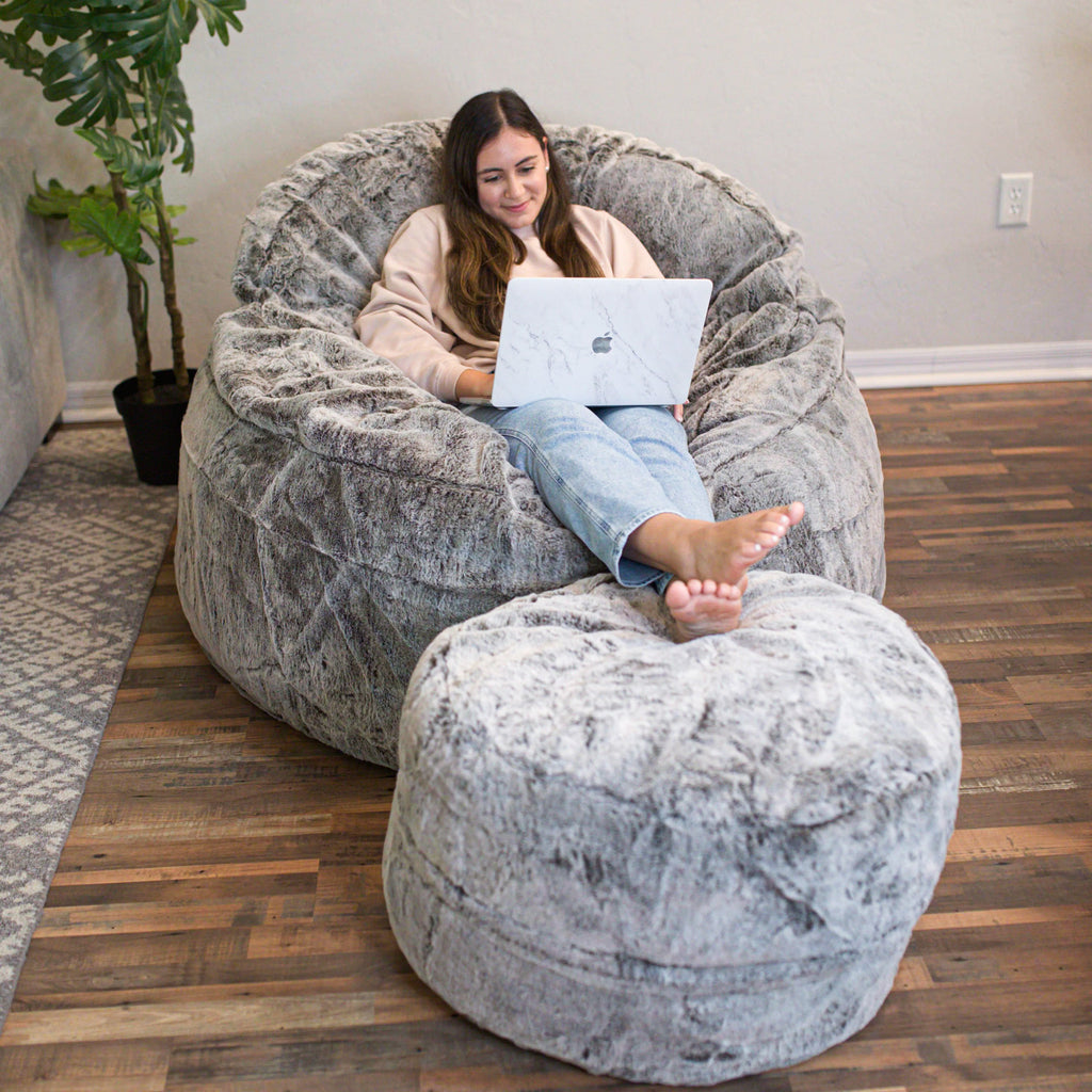 Woman using a laptop on a large gray bean bag chair in a room with wooden flooring and a plant.