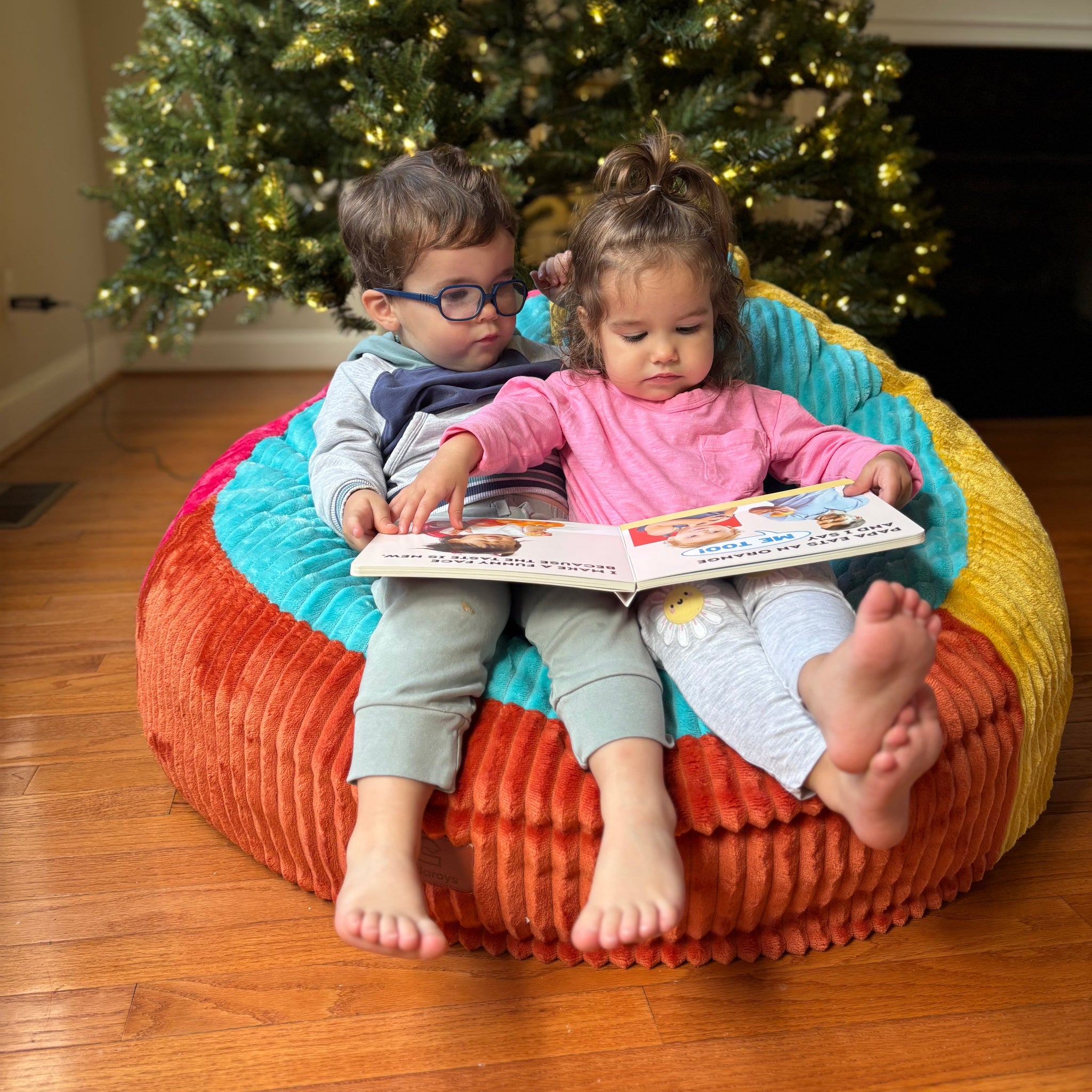 Two children sitting on a colorful bean bag reading a book in front of a Christmas tree.