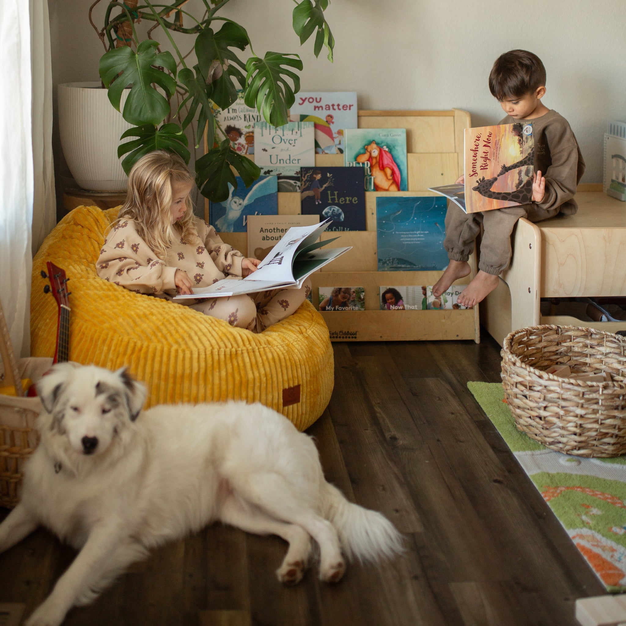 Two children reading books in a cozy room with a dog and a plant.