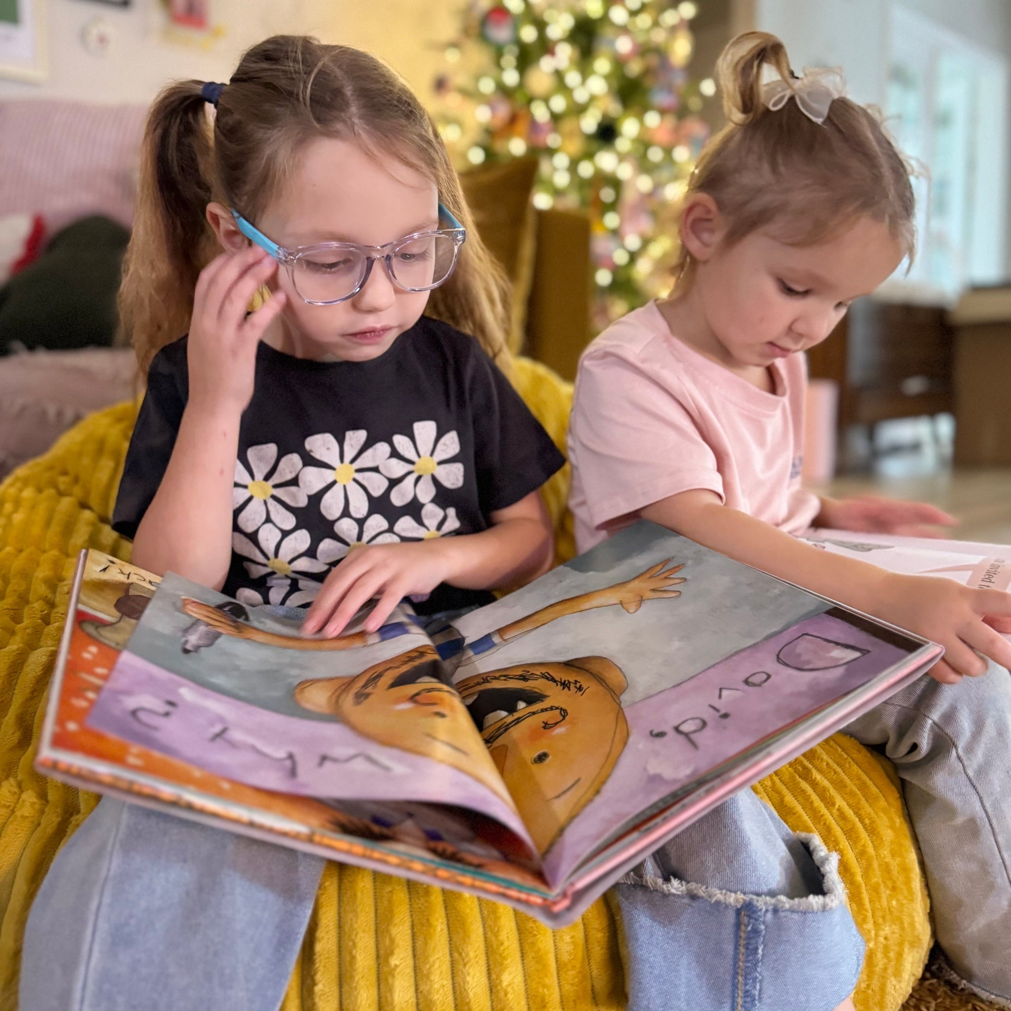 Two young girls reading a book together in a cozy living room with a Christmas tree in the background.