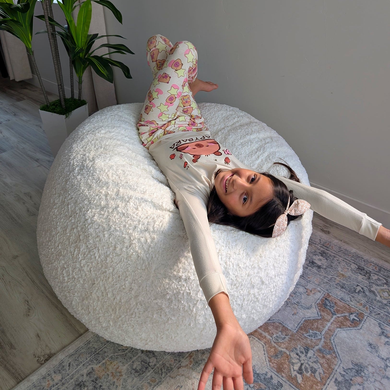 Child lying on a large white bean bag chair in a room with a plant and rug.