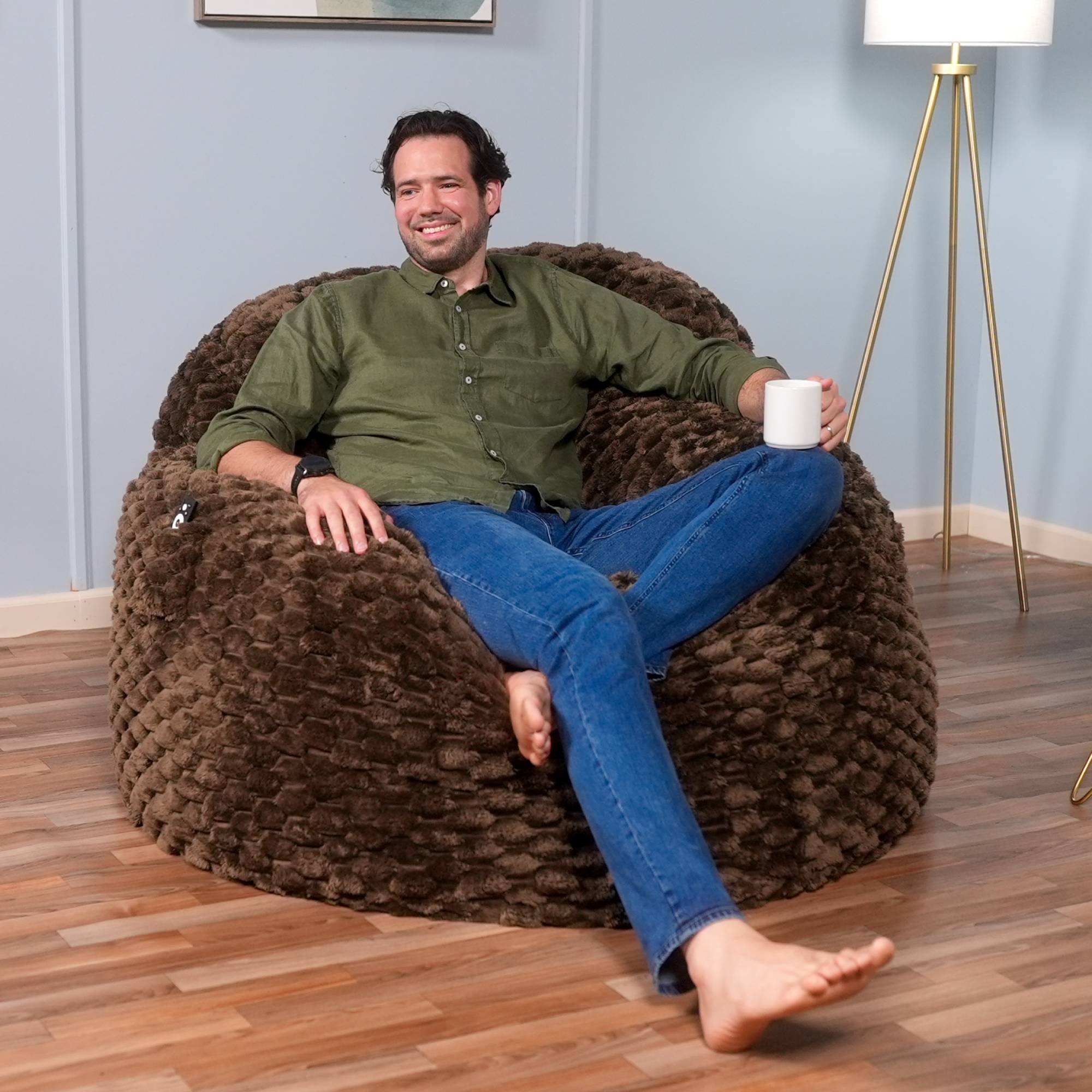 Man sitting on a brown textured bean bag chair holding a white mug in a room with a lamp and wall art.