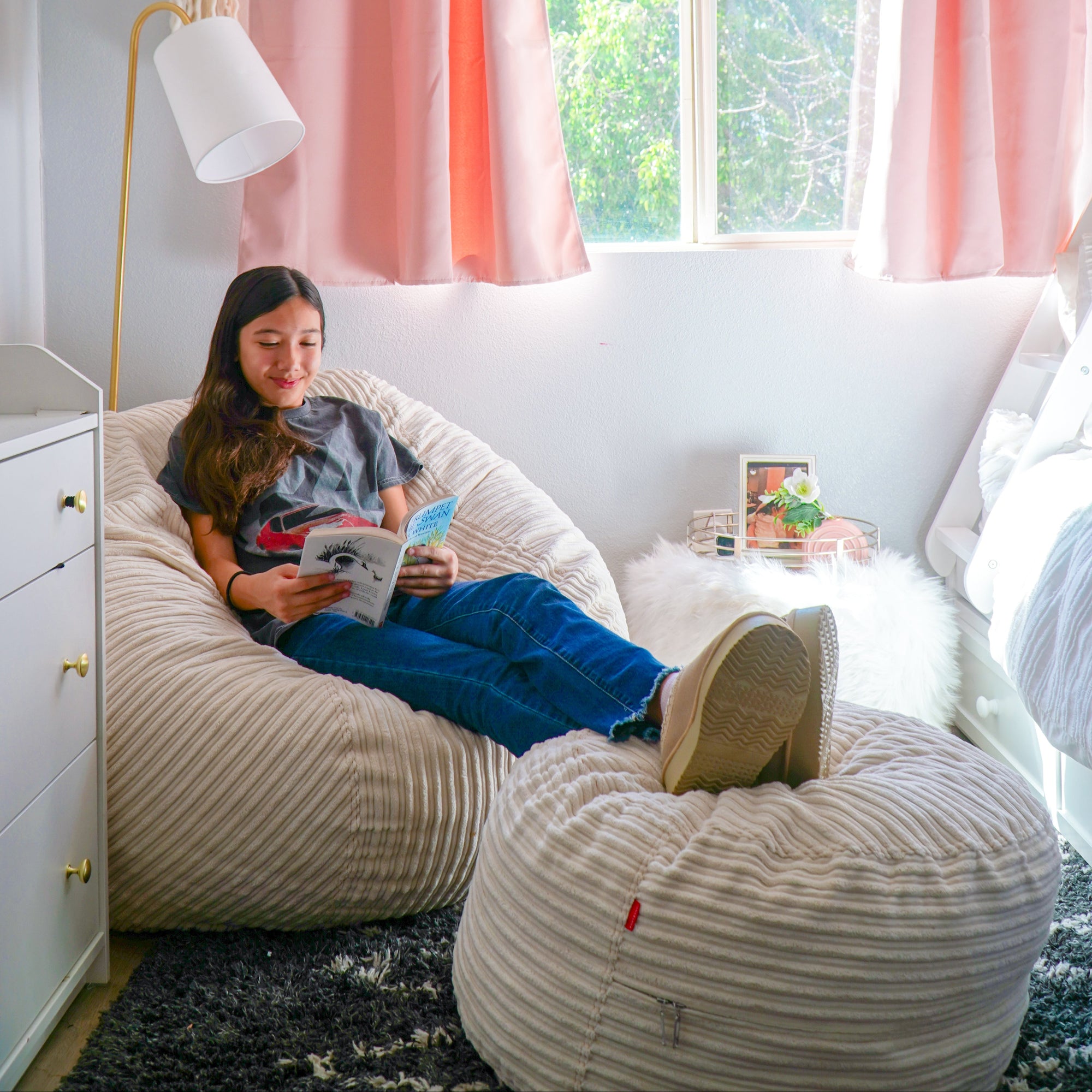 Girl reading a book on a bean bag chair in a room with pink curtains and a window.