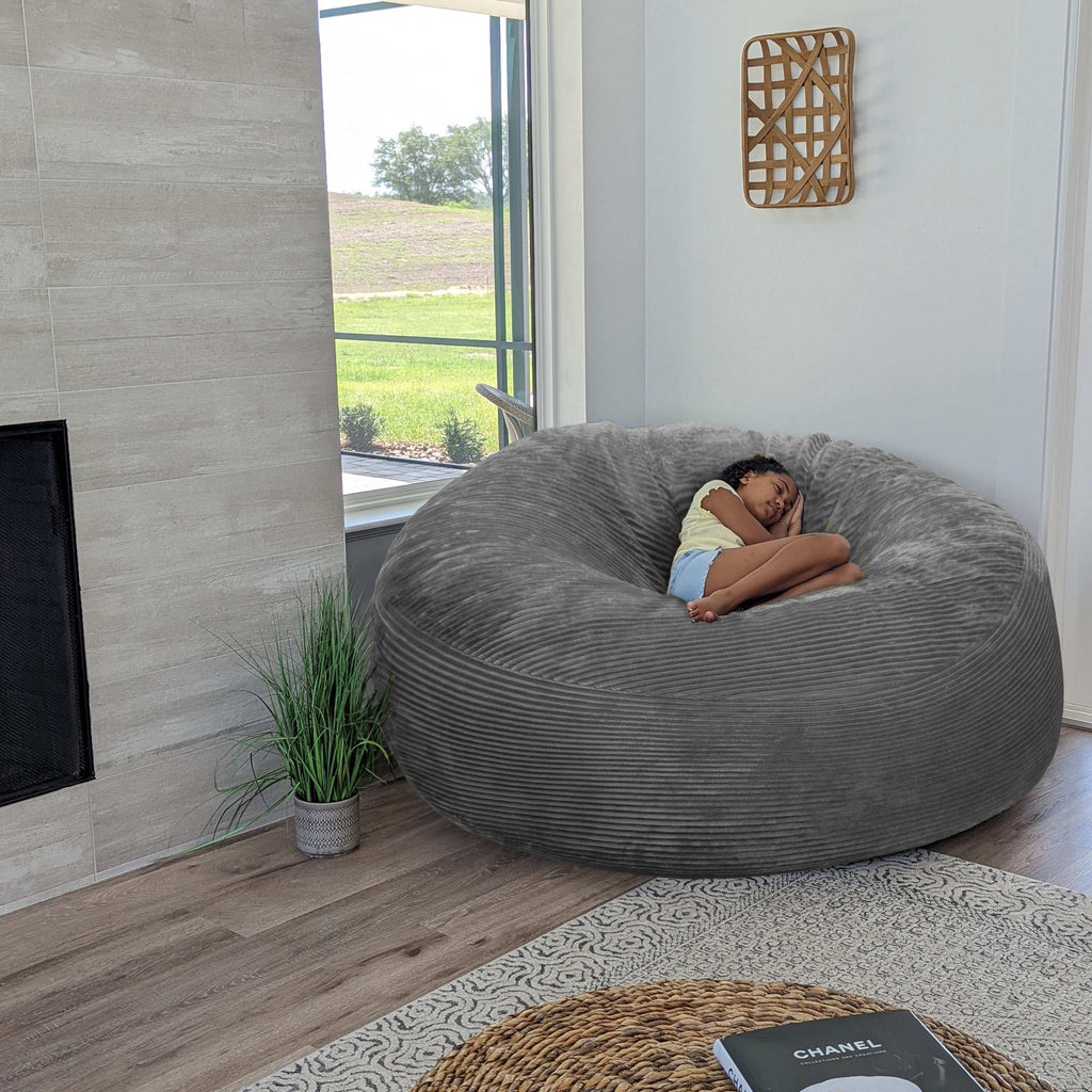 A girl sits on a cordaroys plush corduroy giant bean bag chair in gray. 