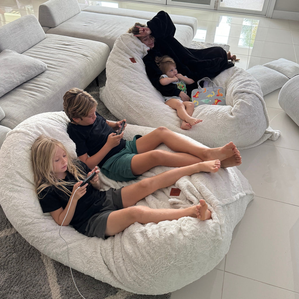 Four kids sits on two cordaroys giant faux fur pillow longers in a living room. 