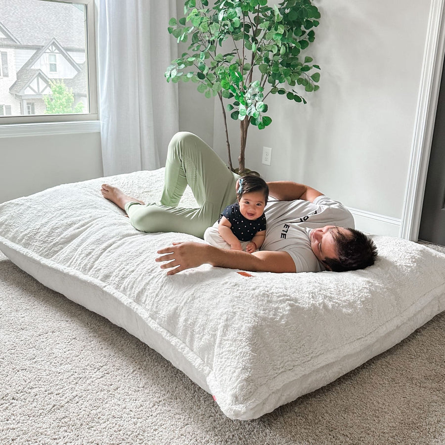 A dad and his child lie on a cordaroys giant pillow lounger flattened out. 