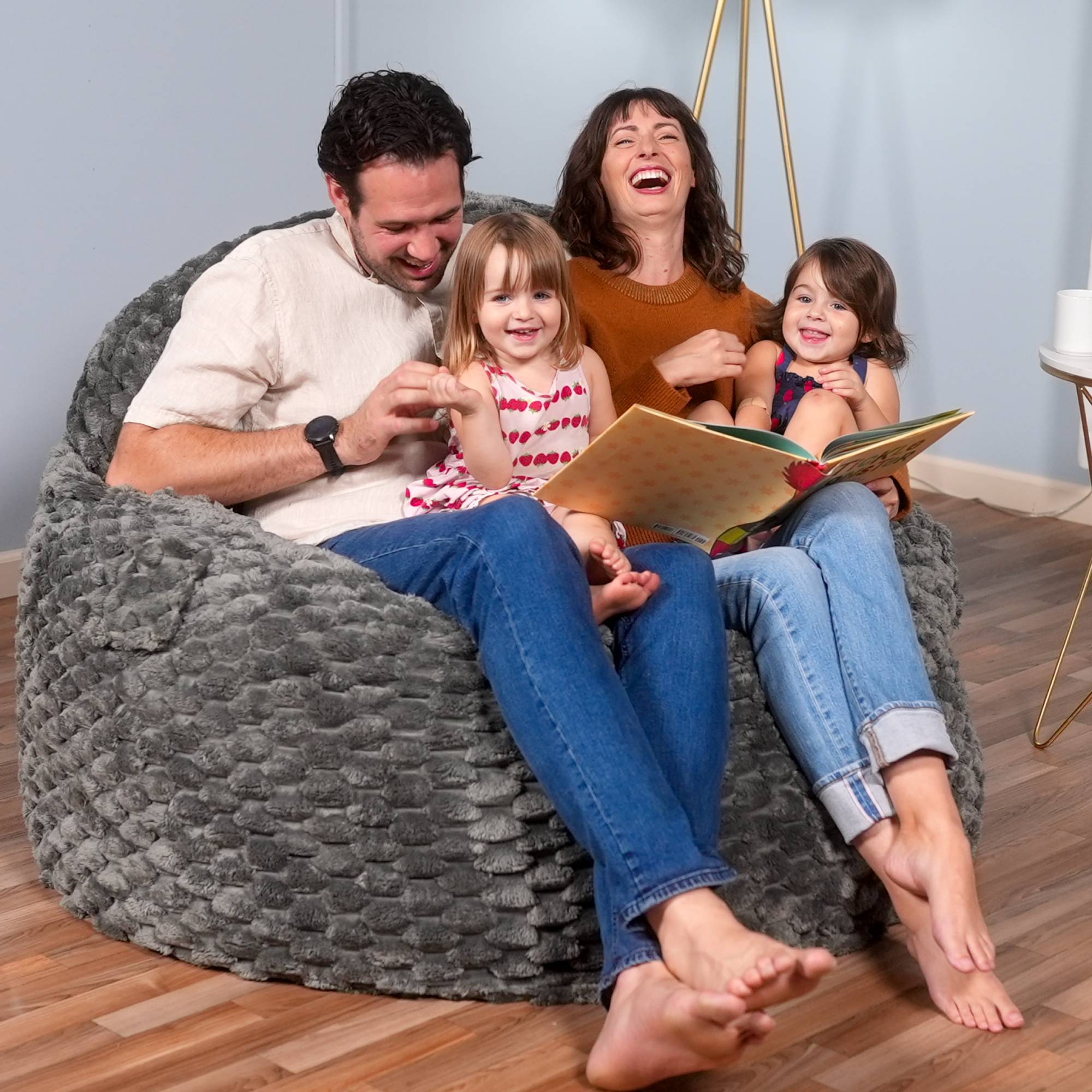 Family of four sitting on a textured gray bean bag chair, reading a book together.