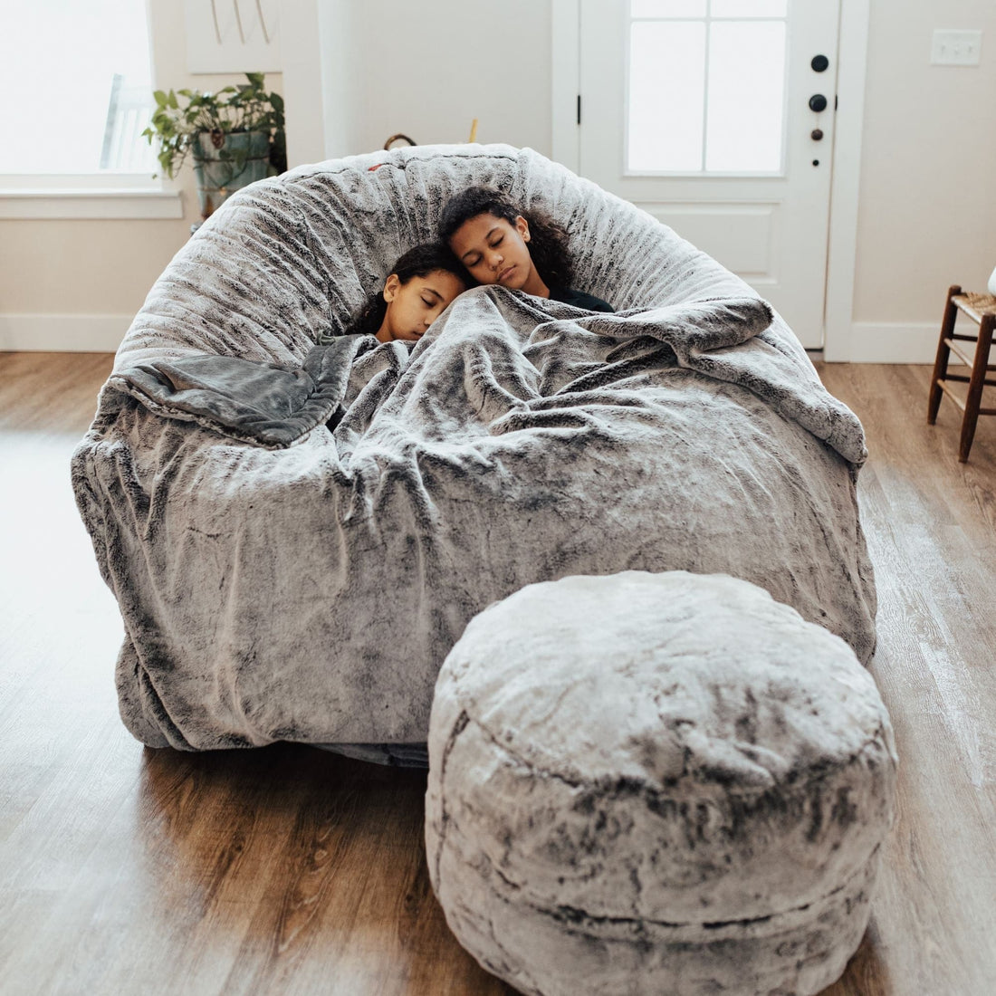 Two kids sit in a cordaroys king chinchilla chair with a footstool wrapped in a blanket. 