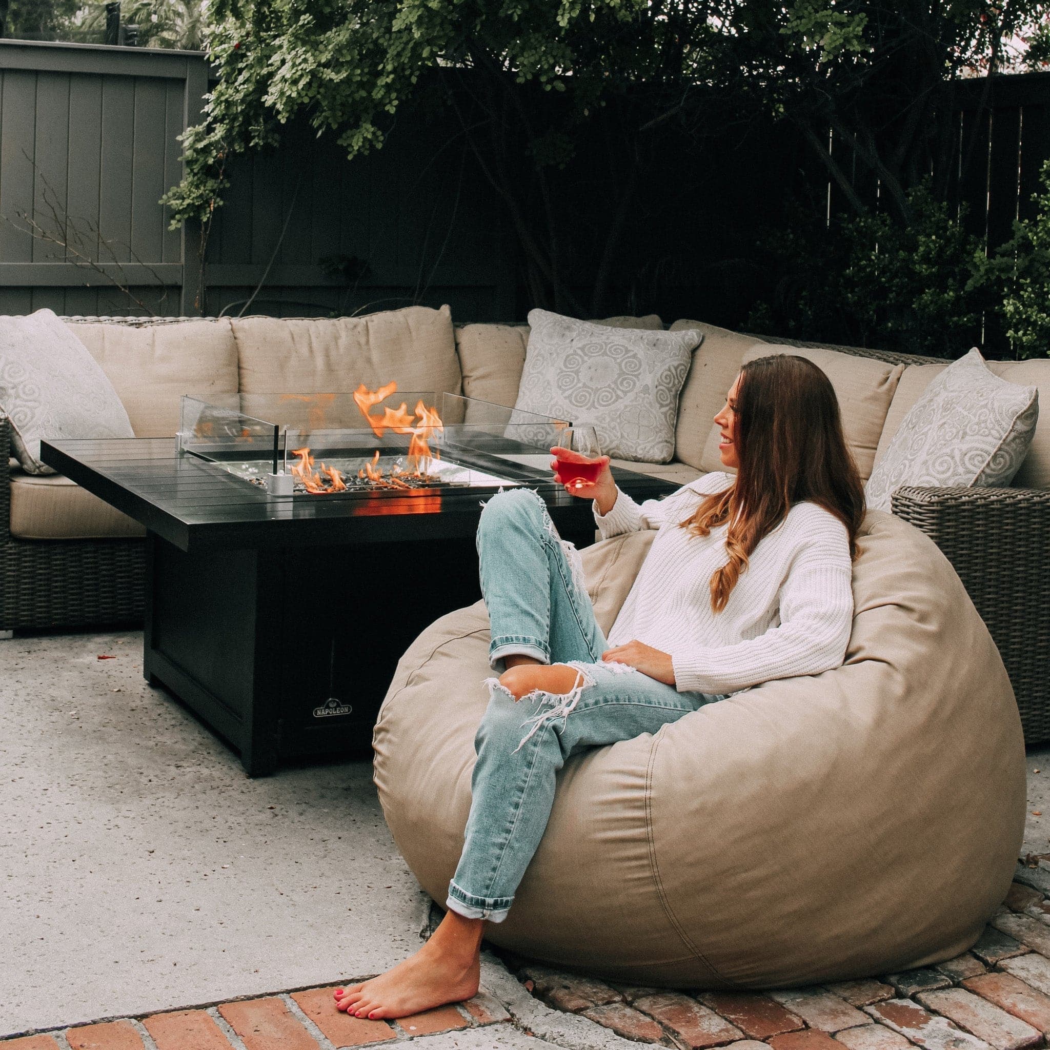 A woman sits in a tan outdoor bean bag by cordaroys next to a fire pit. 