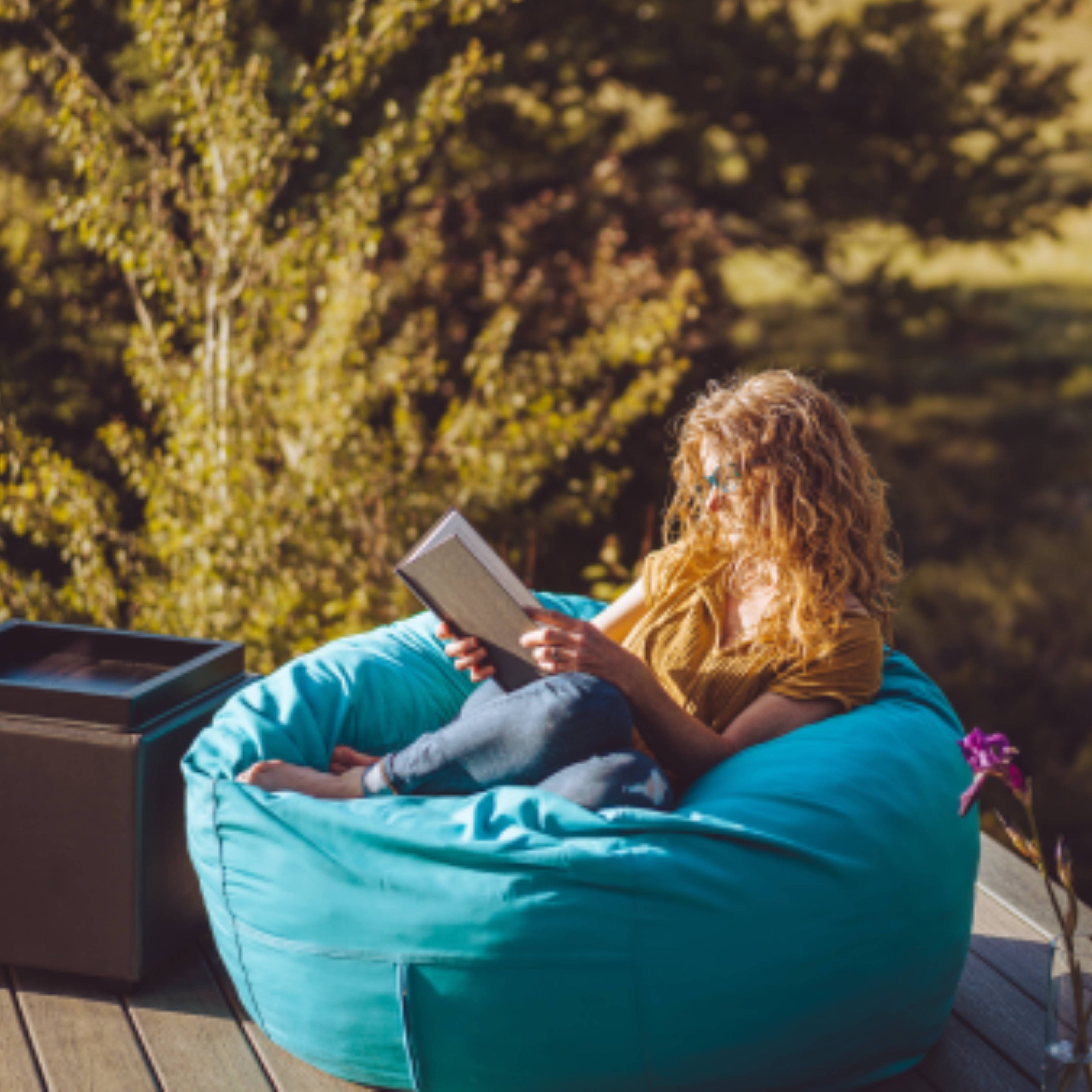 A woman reads a book outside on a cordaroys outdoor bean bag. 