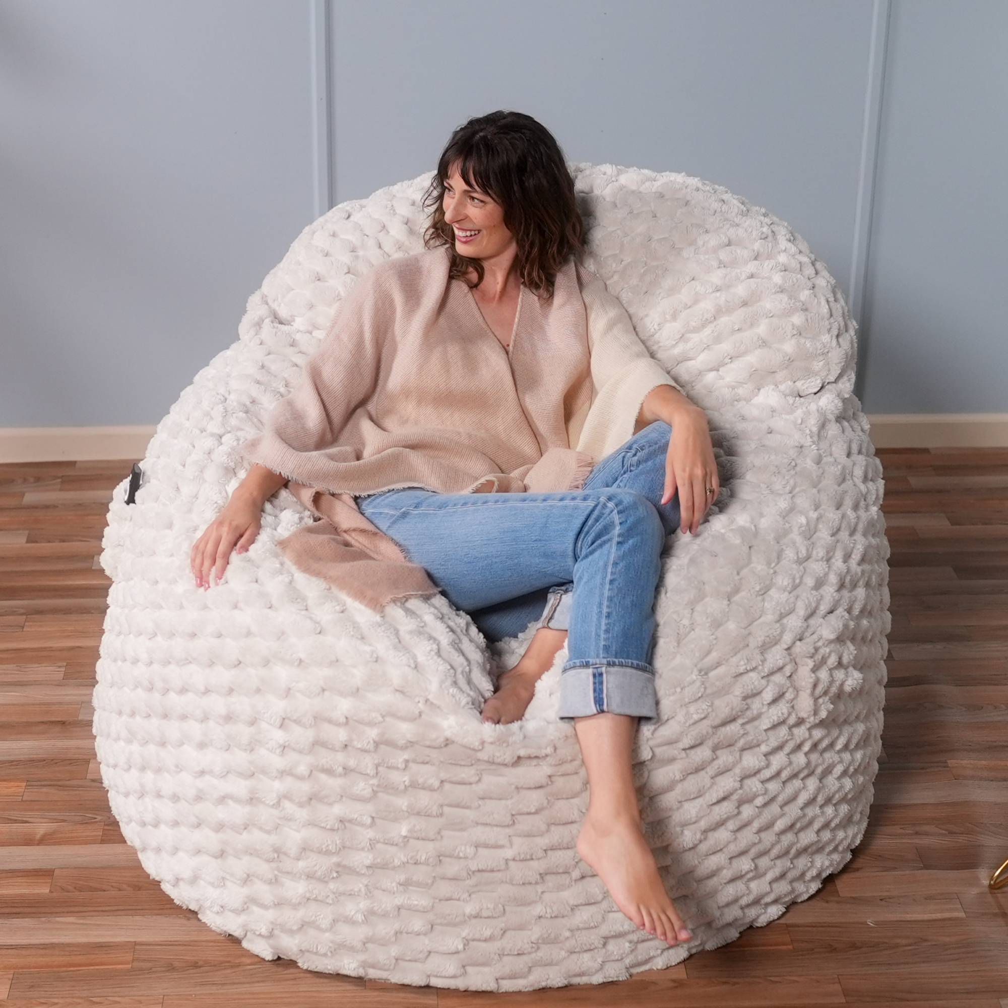 Woman sitting on a textured white bean bag chair in a room with wooden flooring and a gray wall.