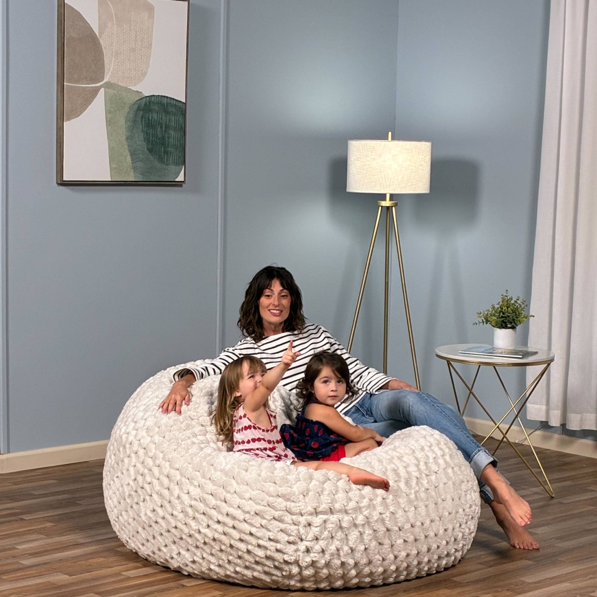 Woman and two children sitting on a large white bean bag chair in a room with a lamp and table.